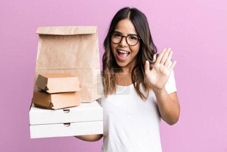 Téléchargez les photos : Hispanic pretty woman smiling happily, waving hand, welcoming and greeting you. delivery and take away fast food concept - en image libre de droit