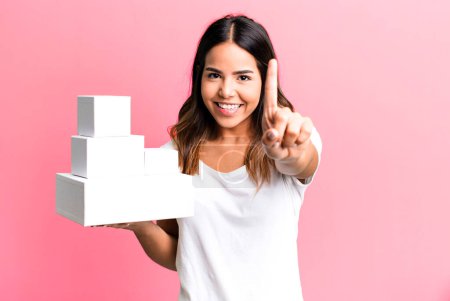 Photo for Hispanic pretty woman smiling proudly and confidently making number one with blank packages boxes - Royalty Free Image
