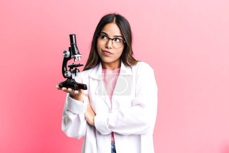 Photo for Hispanic pretty woman shrugging, feeling confused and uncertain. scients student with a microscope - Royalty Free Image