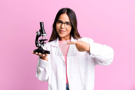 Photo for Hispanic pretty woman smiling cheerfully, feeling happy and pointing to the side. scients student with a microscope - Royalty Free Image