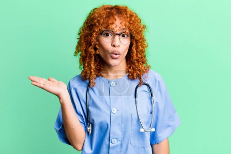 Photo for Red hair pretty woman looking surprised and shocked, with jaw dropped holding an object. nurse concept - Royalty Free Image