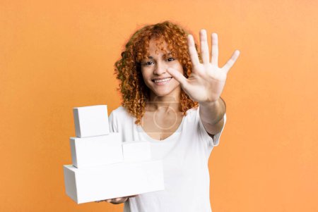 Foto de Cabello rojo bonita mujer sonriendo y buscando amigable, mostrando el número cinco. concepto cajas de paquetes en blanco - Imagen libre de derechos