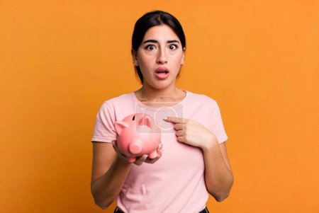 Photo for Looking shocked and surprised with mouth wide open, pointing to self. with a piggy bank - Royalty Free Image