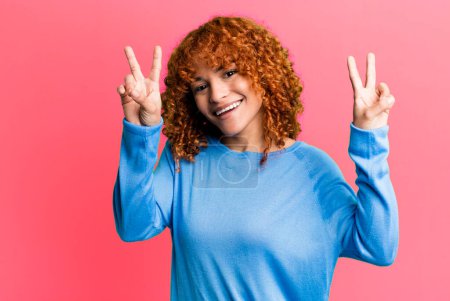Téléchargez les photos : Redhair pretty woman smiling and looking happy, friendly and satisfied, gesturing victory or peace with both hands - en image libre de droit