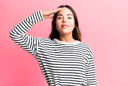 Photo for Hispanic pretty woman greeting the camera with a military salute in an act of honor and patriotism, showing respect - Royalty Free Image