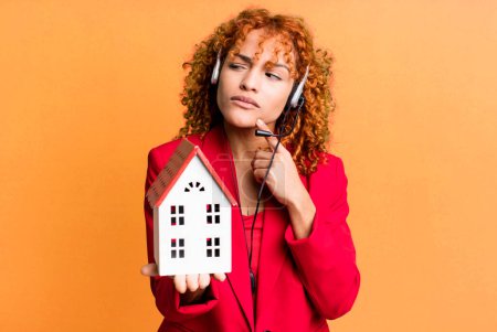 Foto de Pelo rojo bonita mujer agente de bienes raíces con un modelo de casa y auriculares - Imagen libre de derechos