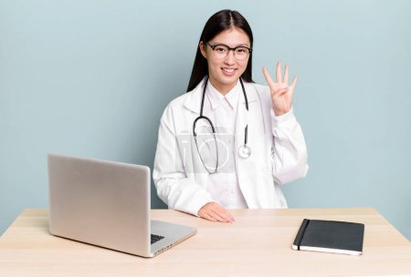 Foto de Pretty asian woman smiling and looking friendly, showing number four. physician desk and laptop - Imagen libre de derechos