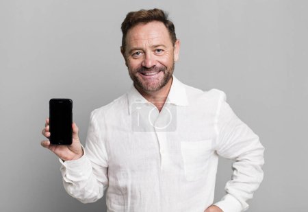 Foto de Middle age man smiling happily with a hand on hip and confident. showing a smartphone - Imagen libre de derechos