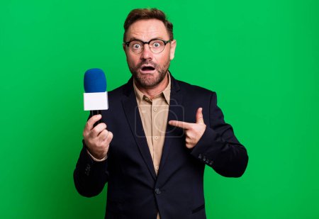 Photo for Middle age man looking shocked and surprised with mouth wide open, pointing to self. journalist and a microphone concept - Royalty Free Image