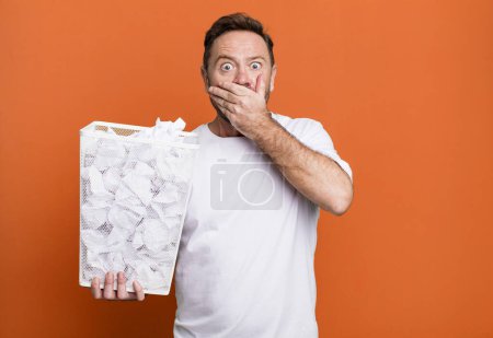 Foto de Middle age man covering mouth with hands with a shocked. paper balls trash basket - Imagen libre de derechos