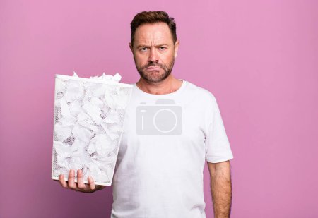 Foto de Middle age man feeling sad and whiney with an unhappy look and crying. paper balls trash basket - Imagen libre de derechos