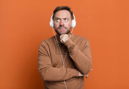 Téléchargez les photos : Middle age man thinking, feeling doubtful and confused. listening music with a headphones - en image libre de droit