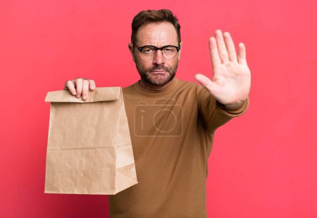 Téléchargez les photos : Middle age man looking serious showing open palm making stop gesture. take away paper bag - en image libre de droit