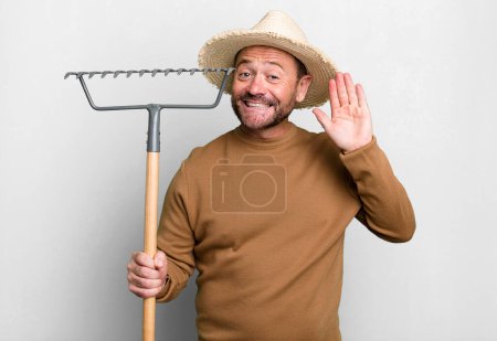 Photo for Middle age man smiling happily, waving hand, welcoming and greeting you. farmer with a rake - Royalty Free Image
