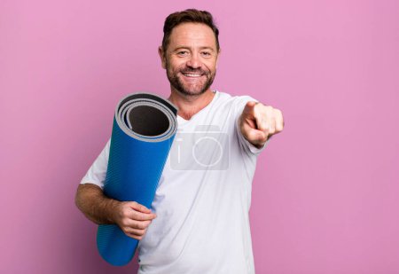 Foto de Middle age man pointing at camera choosing you. with a yoga matt. fitness concept - Imagen libre de derechos