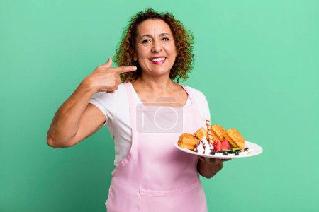 Foto de Pretty middle age woman smiling confidently pointing to own broad smile. home made waffles concept - Imagen libre de derechos