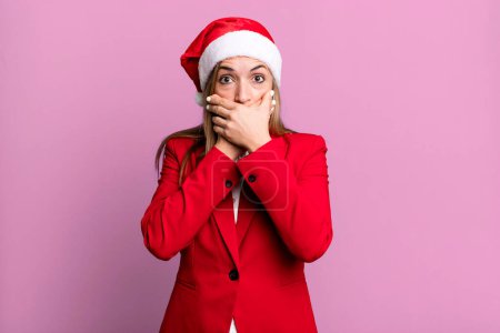 Foto de Pretty blonde woman covering mouth with hands with a shocked. christmas and santa hat concept - Imagen libre de derechos