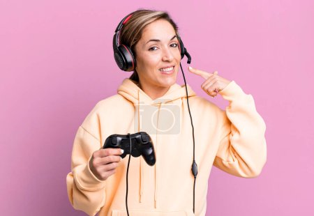 Photo for Pretty blonde woman smiling confidently pointing to own broad smile. gamer with headset and a controller - Royalty Free Image