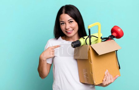 Foto de Mujer bonita hispana mirando emocionada y sorprendida señalando a un lado con una caja de herramientas - Imagen libre de derechos