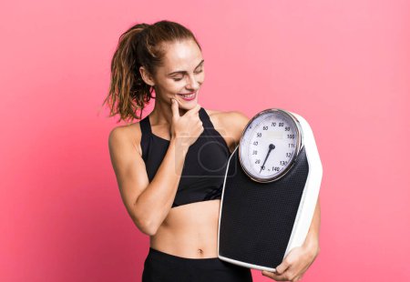 Téléchargez les photos : Hispanic pretty woman smiling with a happy, confident expression with hand on chin. fitness, diet and weight scale concept - en image libre de droit