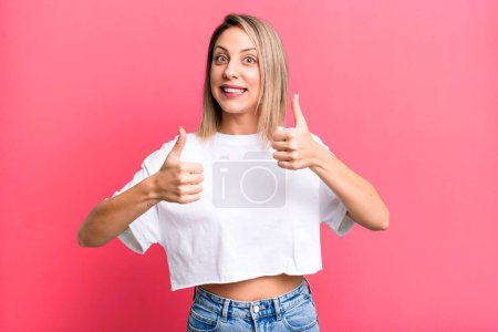Photo for Blonde adult woman smiling broadly looking happy, positive, confident and successful, with both thumbs up - Royalty Free Image