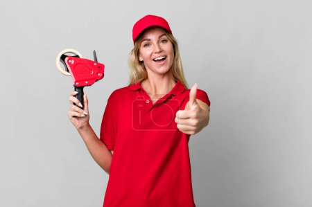 Photo for Caucasian blonde woman feeling proud,smiling positively with thumbs up. shipping packer concept - Royalty Free Image