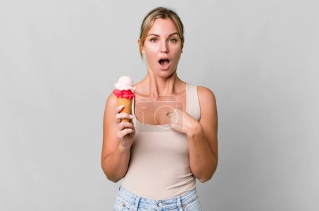 Foto de Caucasian blonde woman looking shocked and surprised with mouth wide open, pointing to self. ice cream concept - Imagen libre de derechos