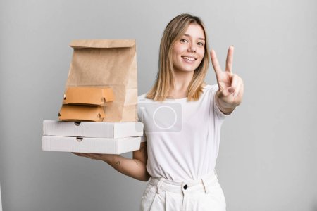 Foto de Joven bonita mujer sonriendo y buscando amigable, mostrando el número dos. entrega y comida para llevar concepto - Imagen libre de derechos