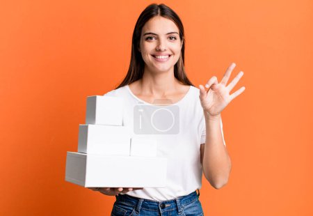 Photo for Young pretty woman feeling happy, showing approval with okay gesture. blank white boxes - Royalty Free Image