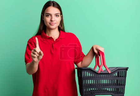 Téléchargez les photos : Young pretty woman smiling proudly and confidently making number one. empty shopping basket concept - en image libre de droit