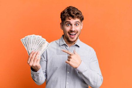 Foto de Young adult caucasian man looking excited and surprised pointing to the side. dollar bank notes concept - Imagen libre de derechos