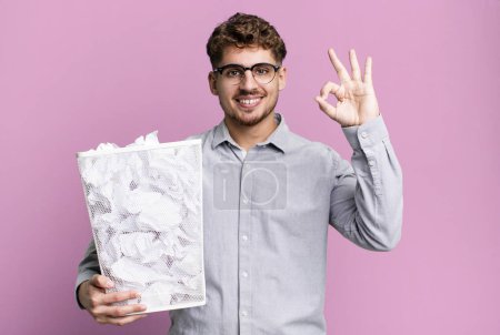 Foto de Young adult caucasian man feeling happy, showing approval with okay gesture with a paper balls trash concept - Imagen libre de derechos