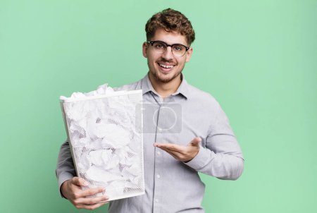 Téléchargez les photos : Jeune homme caucasien adulte souriant joyeusement, se sentant heureux et montrant un concept avec un concept de poubelle boules de papier - en image libre de droit