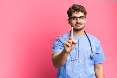 Téléchargez les photos : Jeune homme caucasien adulte souriant et regardant amical, montrant numéro un. concept d'infirmière - en image libre de droit