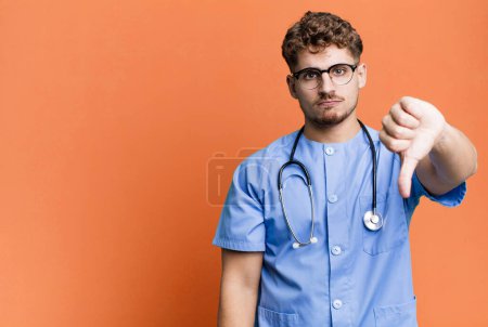 Foto de Young adult caucasian man feeling cross,showing thumbs down. nurse concept - Imagen libre de derechos