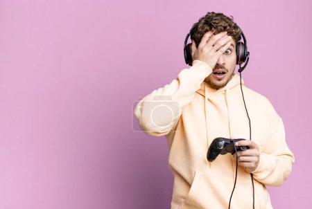 Photo for Young adult caucasian man looking shocked, scared or terrified, covering face with hand with headset and a controller. gamer concept - Royalty Free Image