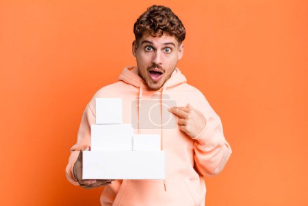 Photo for Young adult caucasian man looking shocked and surprised with mouth wide open, pointing to self. blank different packages concept - Royalty Free Image