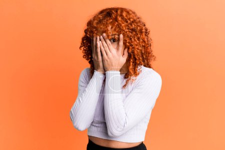 Photo for Redhair pretty woman feeling scared or embarrassed, peeking or spying with eyes half-covered with hands - Royalty Free Image