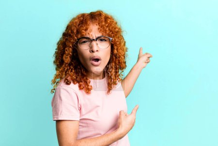 Foto de Redhair pretty woman feeling shocked and surprised, pointing to copy space on the side with amazed, open-mouthed look - Imagen libre de derechos