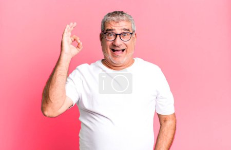 middle age senior man feeling successful and satisfied, smiling with mouth wide open, making okay sign with hand