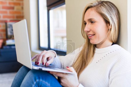Téléchargez les photos : Jeune adulte jolie femme blonde en utilisant son ordinateur portable à la maison - en image libre de droit