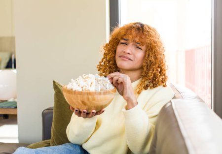 Téléchargez les photos : Young red hair latin pretty woman eating popcorns on a couch and watching a film - en image libre de droit
