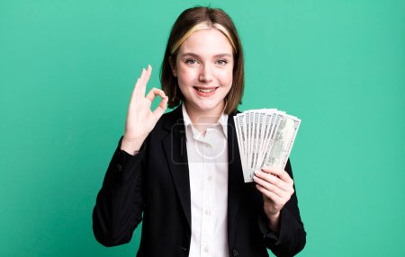 Téléchargez les photos : Young pretty woman feeling happy, showing approval with okay gesture. dollar banknotes - en image libre de droit