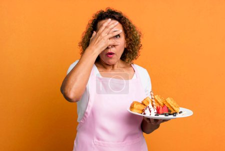 Foto de Pretty middle age woman looking shocked, scared or terrified, covering face with hand. home made waffles concept - Imagen libre de derechos