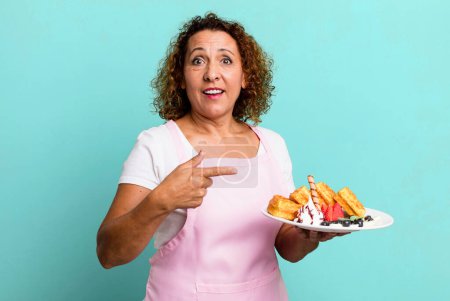 Foto de Pretty middle age woman looking excited and surprised pointing to the side. home made waffles concept - Imagen libre de derechos