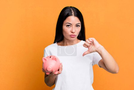 Téléchargez les photos : Hispanic pretty woman feeling cross,showing thumbs down with a piggy bank - en image libre de droit