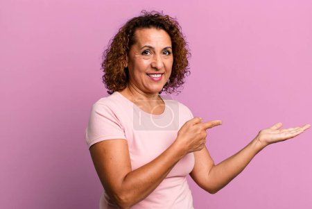 Foto de Mujer hispana de mediana edad sonriendo alegremente y apuntando a copiar espacio en la palma de la mano, mostrando o publicitando un objeto - Imagen libre de derechos
