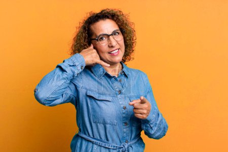 Téléchargez les photos : Femme hispanique d'âge moyen souriant joyeusement et pointant vers la caméra tout en faisant un appel vous geste plus tard, parler au téléphone - en image libre de droit