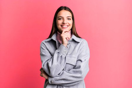 Photo for Pretty young adult woman looking happy and smiling with hand on chin, wondering or asking a question, comparing options - Royalty Free Image