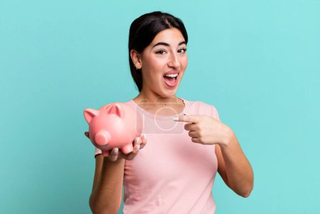 Foto de Looking excited and surprised pointing to the side. with a piggy bank - Imagen libre de derechos